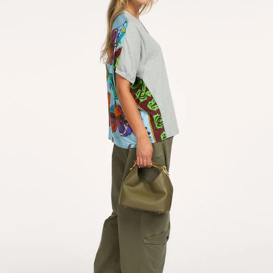 A woman modeling a light gray oversized t-shirt with a colorful abstract print on the back, paired with olive green cargo pants and holding an olive green handbag against a plain white background.
