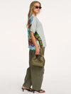 A woman modeling a light gray oversized t-shirt with a colorful abstract print on the back, paired with olive green cargo pants and holding an olive green handbag against a plain white background.