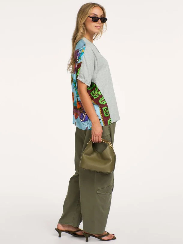 A woman modeling a light gray oversized t-shirt with a colorful abstract print on the back, paired with olive green cargo pants and holding an olive green handbag against a plain white background.