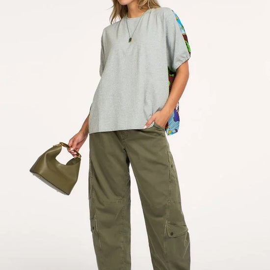 A woman modeling a light gray oversized short-sleeve top with colorful patterned side panels, olive green wide-leg cargo pants, sunglasses, and a small olive green handbag against a plain white studio background.
