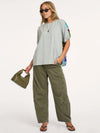 A woman modeling a light gray oversized short-sleeve top with colorful patterned side panels, olive green wide-leg cargo pants, sunglasses, and a small olive green handbag against a plain white studio background.