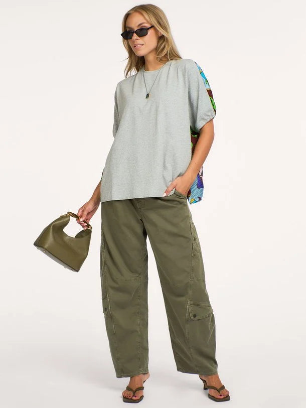 A woman modeling a light gray oversized short-sleeve top with colorful patterned side panels, olive green wide-leg cargo pants, sunglasses, and a small olive green handbag against a plain white studio background.