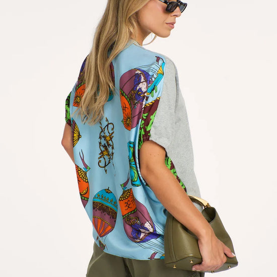 A woman modeling a light blue short-sleeve top with a colorful abstract print on the front and solid light gray on the sleeves, paired with olive green pants and holding an olive green handbag against a plain white background.