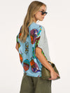 A woman modeling a light blue short-sleeve top with a colorful abstract print on the front and solid light gray on the sleeves, paired with olive green pants and holding an olive green handbag against a plain white background.