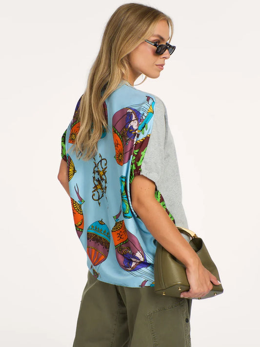 A woman modeling a light blue short-sleeve top with a colorful abstract print on the front and solid light gray on the sleeves, paired with olive green pants and holding an olive green handbag against a plain white background.