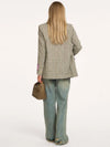 A woman shown from the back wearing a light beige and gray houndstooth blazer, wide-leg light blue jeans, and holding a small woven brown handbag against a plain white background.