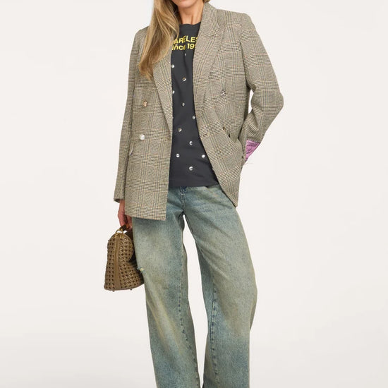 A woman models a double-breasted blazer in a beige and brown houndstooth pattern, styled with a black graphic t-shirt, wide-leg light-wash jeans, and brown shoes, holding a small woven handbag against a plain white studio background.