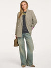 A woman models a double-breasted blazer in a beige and brown houndstooth pattern, styled with a black graphic t-shirt, wide-leg light-wash jeans, and brown shoes, holding a small woven handbag against a plain white studio background.