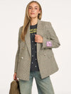 A woman models an oversized double-breasted blazer in a beige and brown plaid pattern, worn over a black graphic t-shirt and light-wash jeans. She is holding a woven brown handbag against a plain off-white studio background.