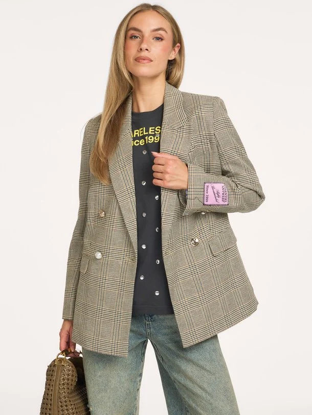 A woman models an oversized double-breasted blazer in a beige and brown plaid pattern, worn over a black graphic t-shirt and light-wash jeans. She is holding a woven brown handbag against a plain off-white studio background.