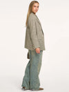 A woman modeling an oversized double-breasted blazer in a muted green and beige houndstooth pattern, paired with wide-leg denim jeans, against a plain white studio background.