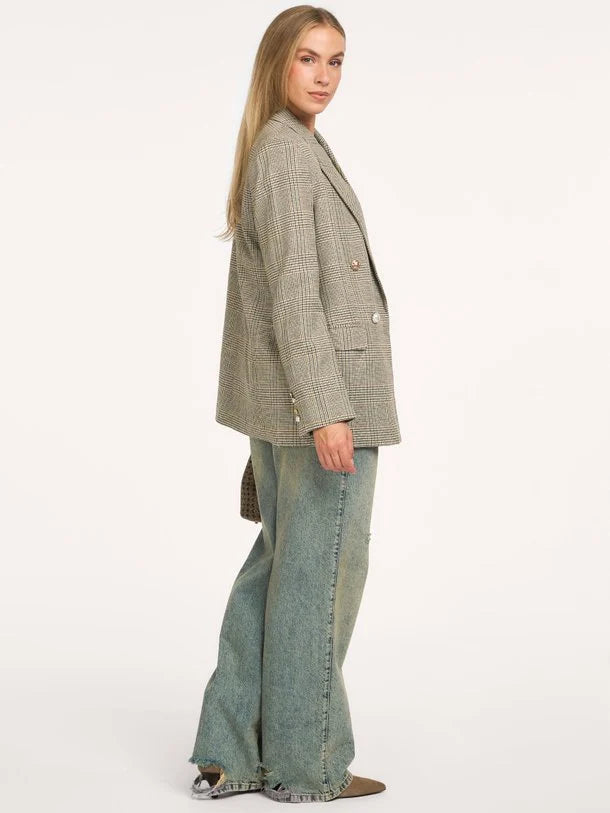 A woman modeling an oversized double-breasted blazer in a muted green and beige houndstooth pattern, paired with wide-leg denim jeans, against a plain white studio background.