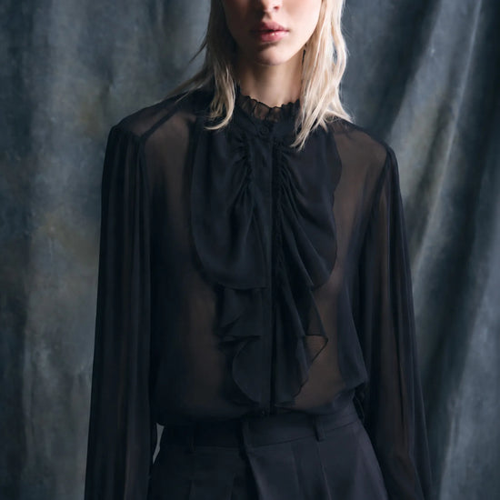 A blonde model wearing a sheer black long-sleeve blouse with a large ruffled bow at the neck, paired with black trousers, set against a dark textured studio backdrop.