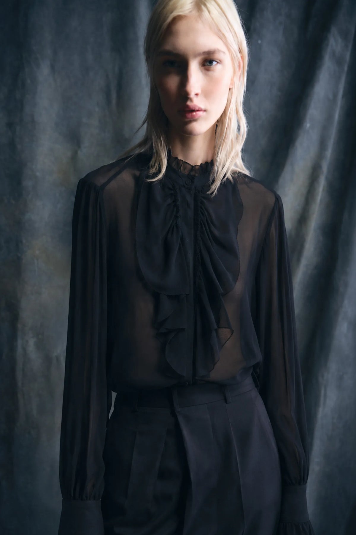 A blonde model wearing a sheer black long-sleeve blouse with a large ruffled bow at the neck, paired with black trousers, set against a dark textured studio backdrop.
