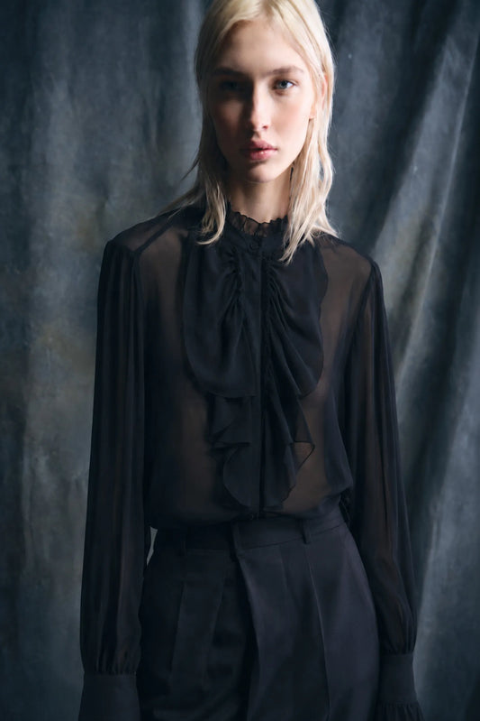 A blonde model wearing a sheer black long-sleeve blouse with a large ruffled bow at the neck, paired with black trousers, set against a dark textured studio backdrop.
