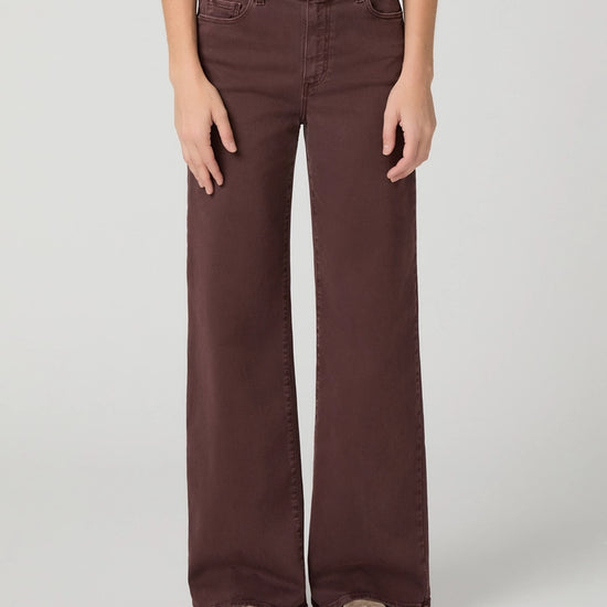 A person wearing dark brown wide-leg jeans with a white top and brown slip-on shoes, standing against a light gray studio background.