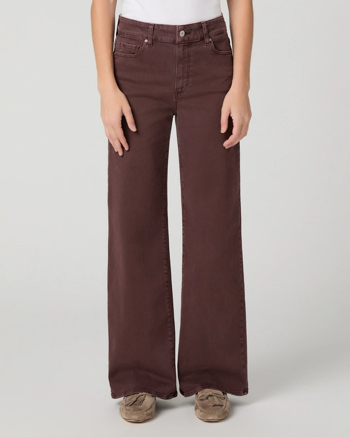 A person wearing dark brown wide-leg jeans with a white top and brown slip-on shoes, standing against a light gray studio background.