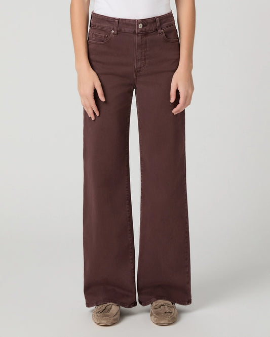 A person wearing dark brown wide-leg jeans with a white top and brown slip-on shoes, standing against a light gray studio background.