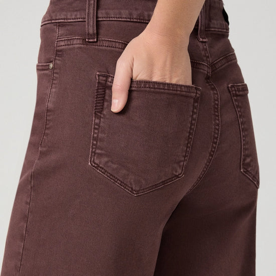 Close-up of a person wearing dark brown denim jeans, showing the back pocket and hand in the pocket, against a light gray background.