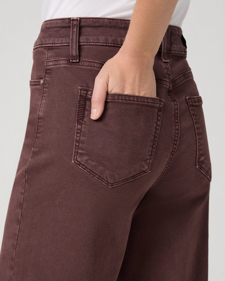 Close-up of a person wearing dark brown denim jeans, showing the back pocket and hand in the pocket, against a light gray background.