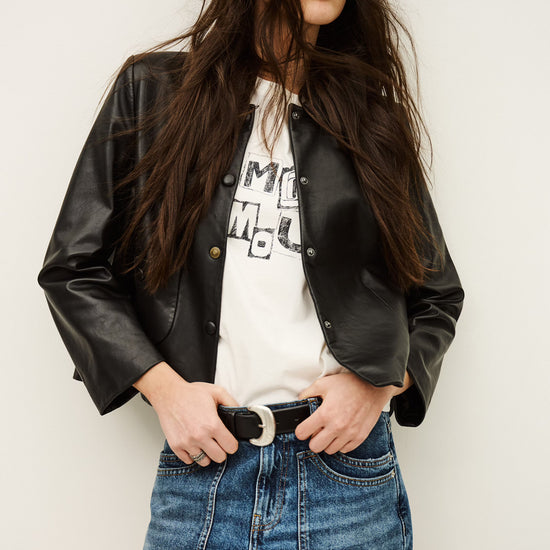 Woman wearing a black leather jacket, white graphic t-shirt, and blue denim shorts against a plain background