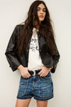 Woman wearing a black leather jacket, white graphic t-shirt, and blue denim shorts against a plain background