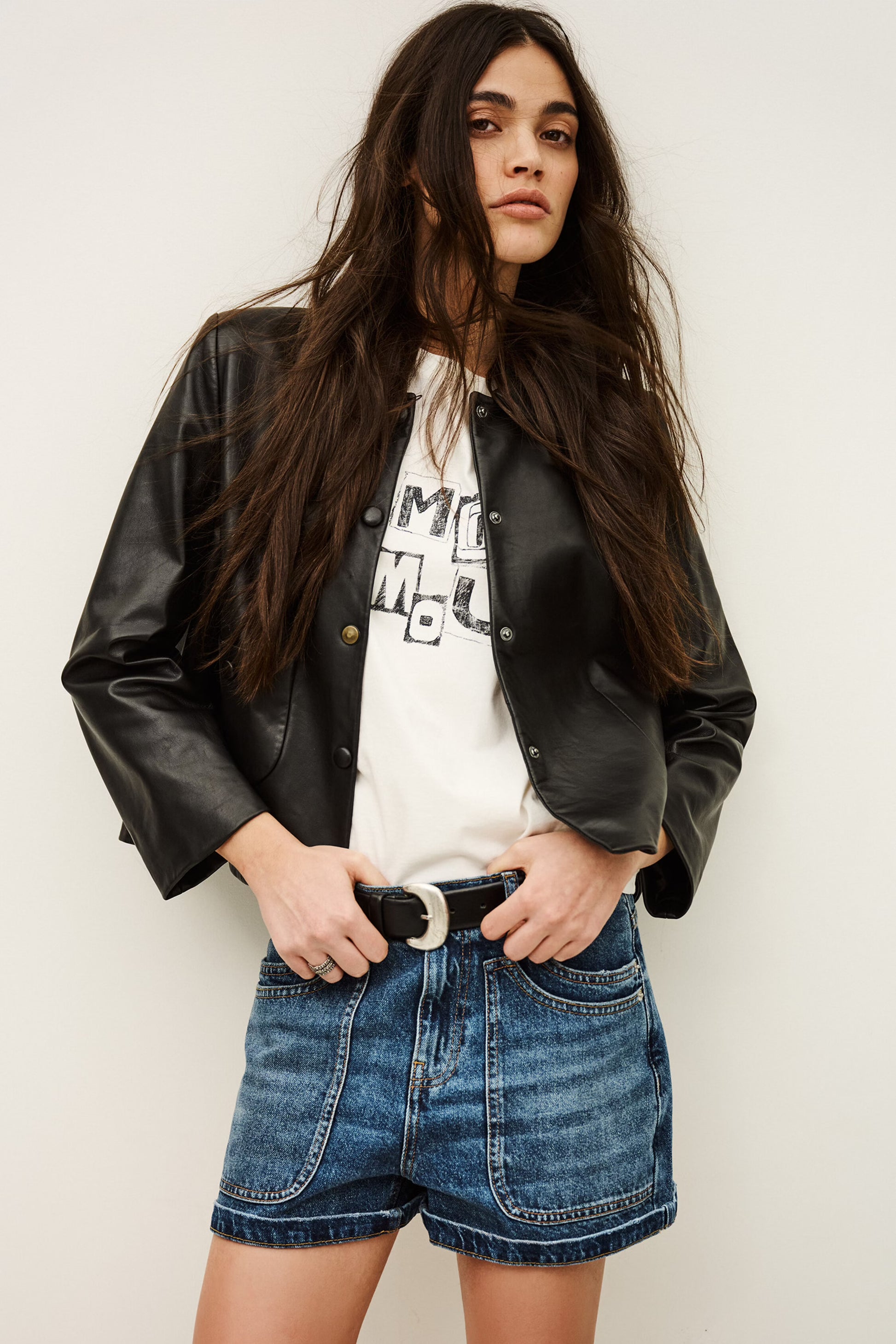 Woman wearing a black leather jacket, white graphic t-shirt, and blue denim shorts against a plain background