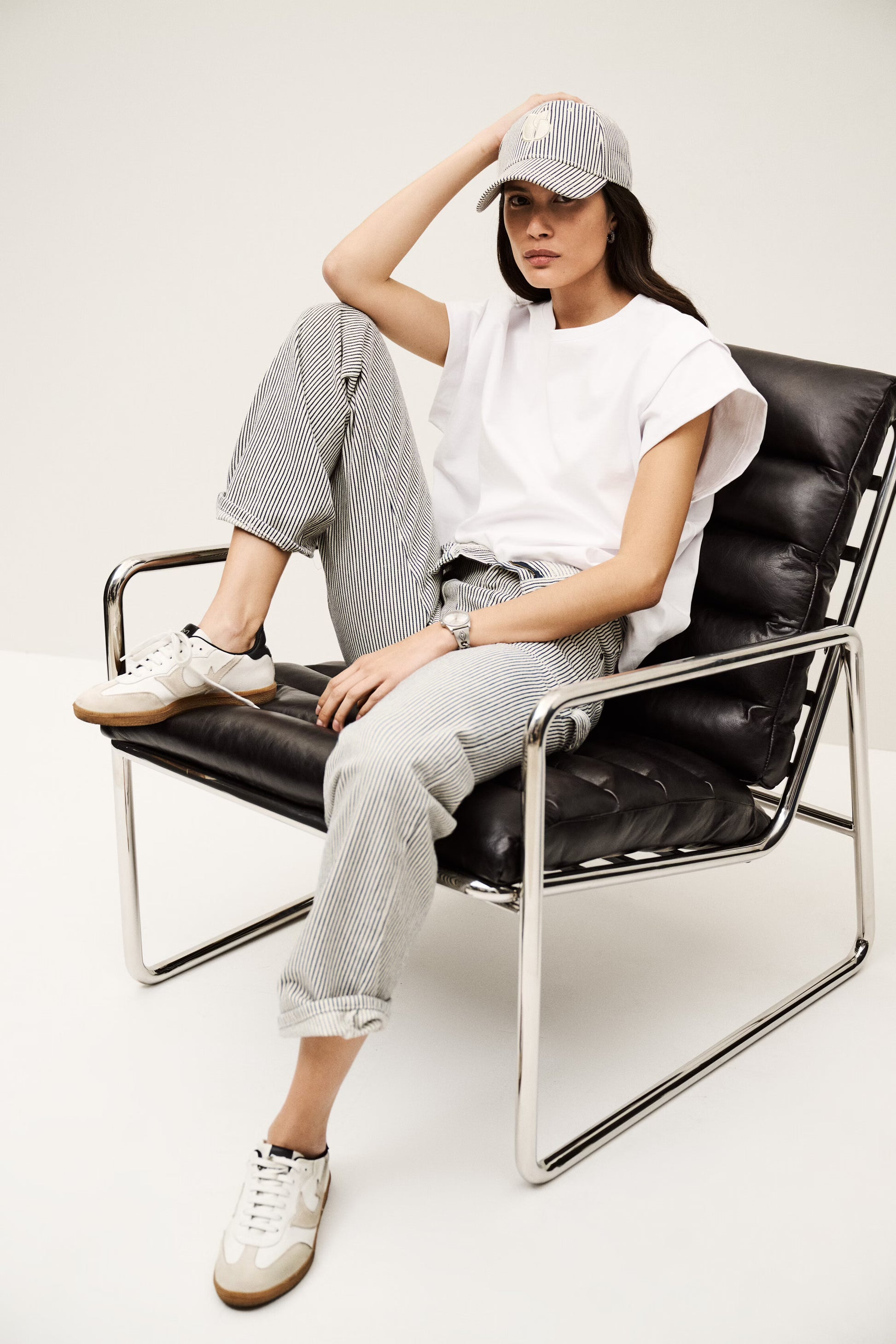 Person sitting on a black chair wearing a white t-shirt, gray pants, and a cap. from ba&sh