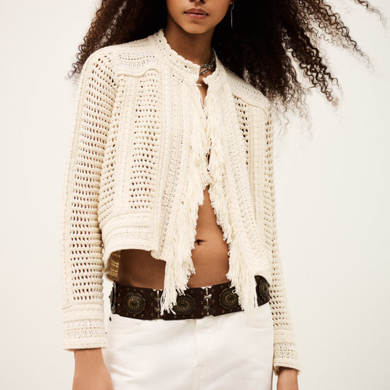 Woman wearing a white crochet cardigan over a black top with white pants on a plain background from bash