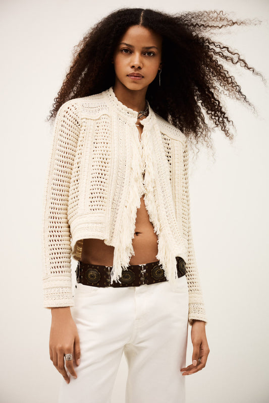 Woman wearing a white crochet cardigan over a black top with white pants on a plain background from bash