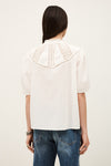 Person wearing a white blouse with lace details and blue jeans on a plain background