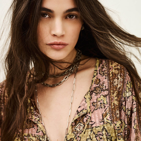 Woman with long hair wearing a patterned top and layered necklaces against a neutral background