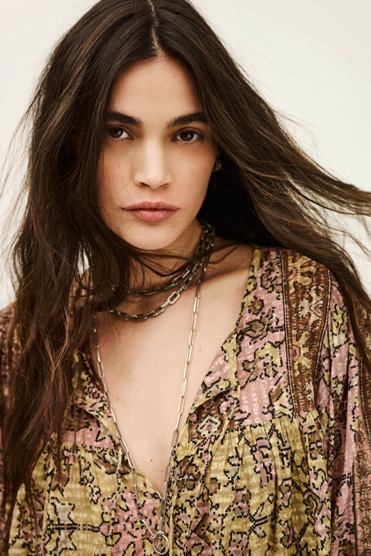 Woman with long hair wearing a patterned top and layered necklaces against a neutral background