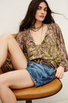 Woman sitting on a chair wearing a patterned blouse and denim shorts.