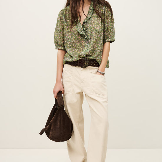 Woman wearing a green patterned blouse and beige pants, holding a brown bag on a white background from ba&sh