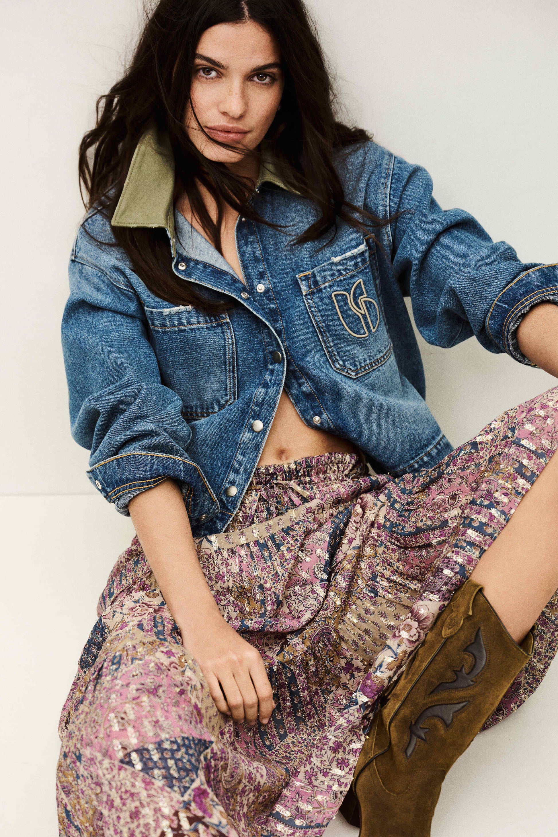 Woman wearing a denim jacket, patterned skirt, and brown boots against a white background