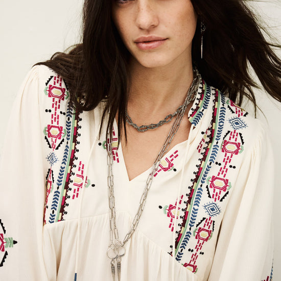 Woman wearing a white blouse with colorful embroidery and layered necklaces against a white background