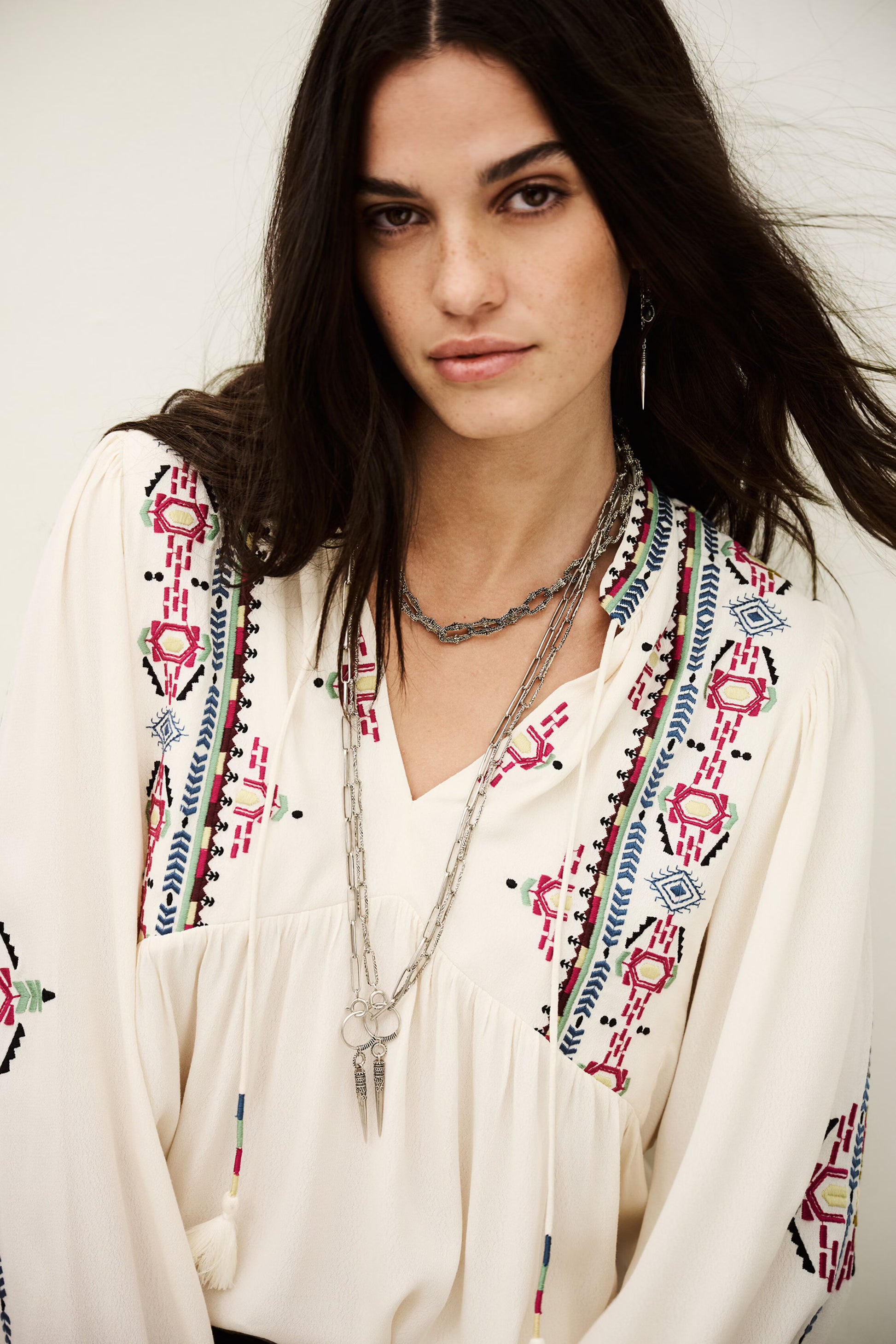 Woman wearing a white blouse with colorful embroidery and layered necklaces against a white background