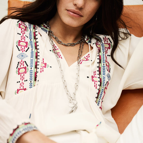 Woman wearing a white embroidered blouse with colorful patterns, sitting on a couch.