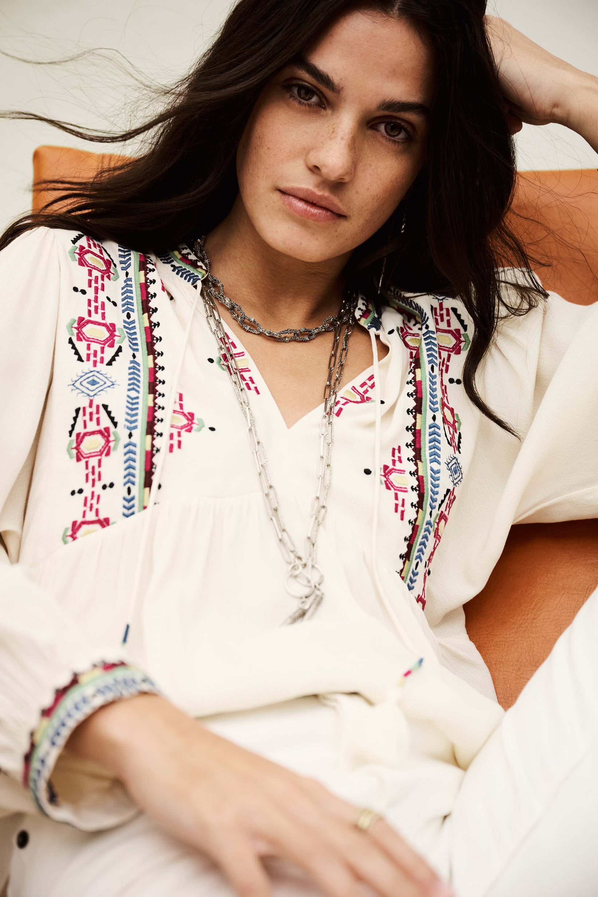 Woman wearing a white embroidered blouse with colorful patterns, sitting on a couch.