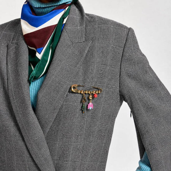 A close-up of a model wearing a gray plaid double-breasted blazer, a blue turtleneck sweater, a striped scarf, and a gold safety-pin style brooch with colorful charms on the blazer chest.