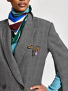 A close-up of a model wearing a gray plaid double-breasted blazer, a blue turtleneck sweater, a striped scarf, and a gold safety-pin style brooch with colorful charms on the blazer chest.