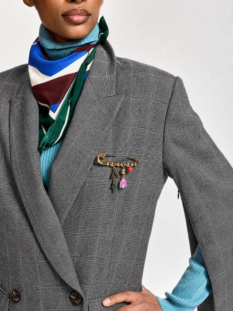 A close-up of a model wearing a gray plaid double-breasted blazer, a blue turtleneck sweater, a striped scarf, and a gold safety-pin style brooch with colorful charms on the blazer chest.