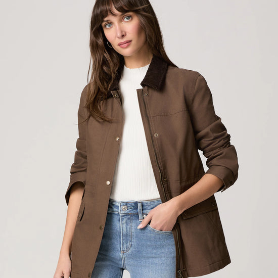 Woman wearing a brown jacket with a white shirt and blue jeans on a light gray background