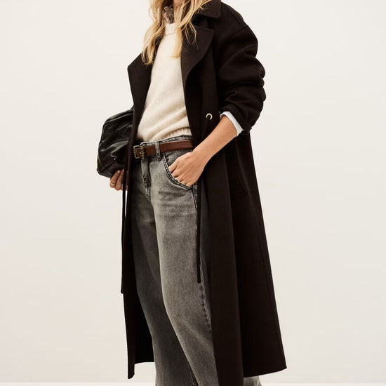 Person wearing a long brown coat, jeans, and a cap on a white background