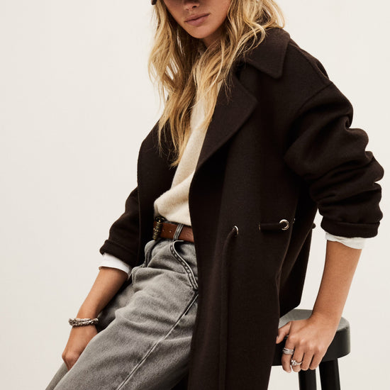 Woman wearing a dark coat, cap, and jeans sitting on a stool against a plain background
