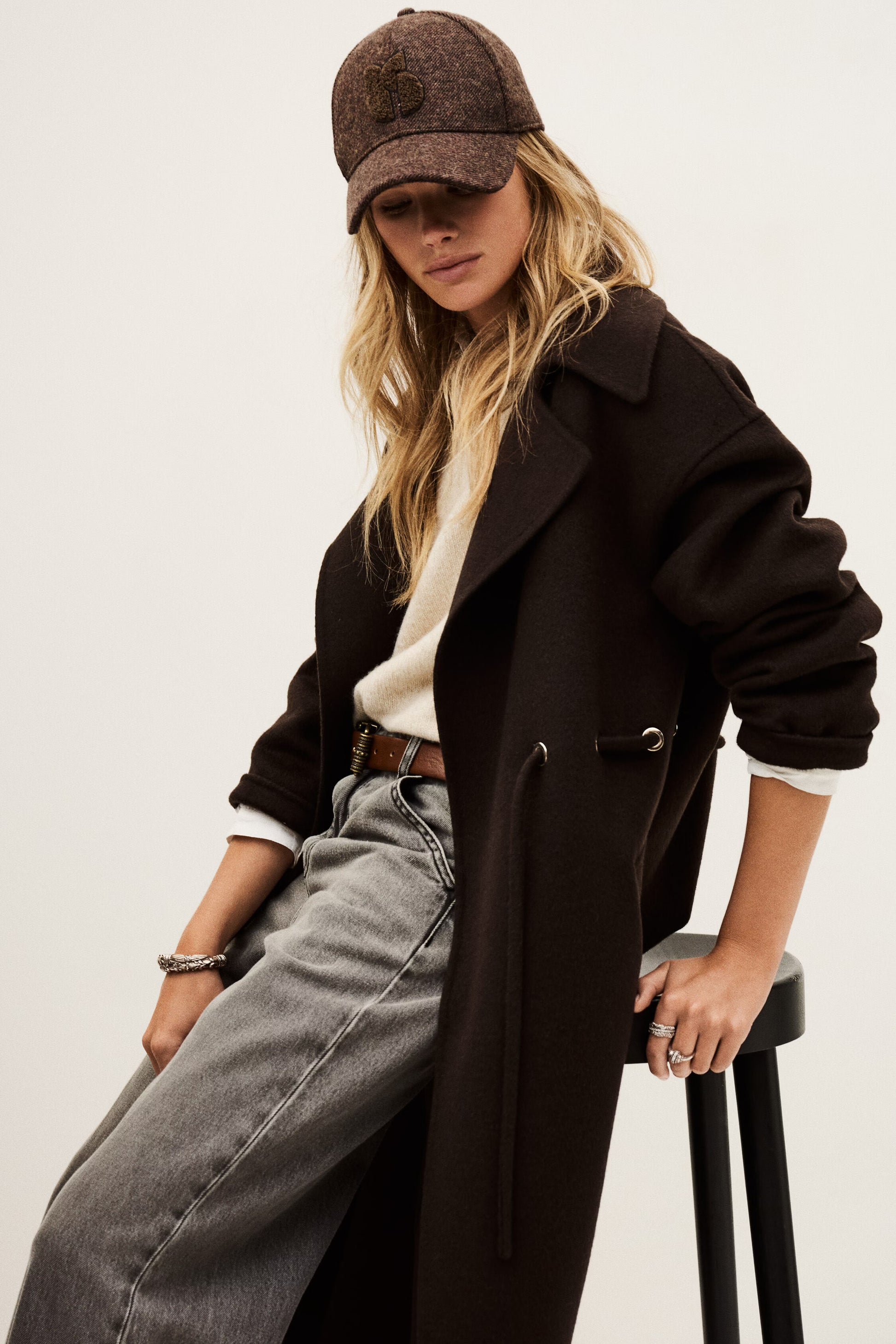 Woman wearing a dark coat, cap, and jeans sitting on a stool against a plain background