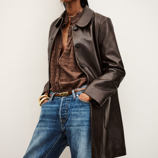 Person wearing a brown leather coat, brown shirt, and blue jeans against a white background