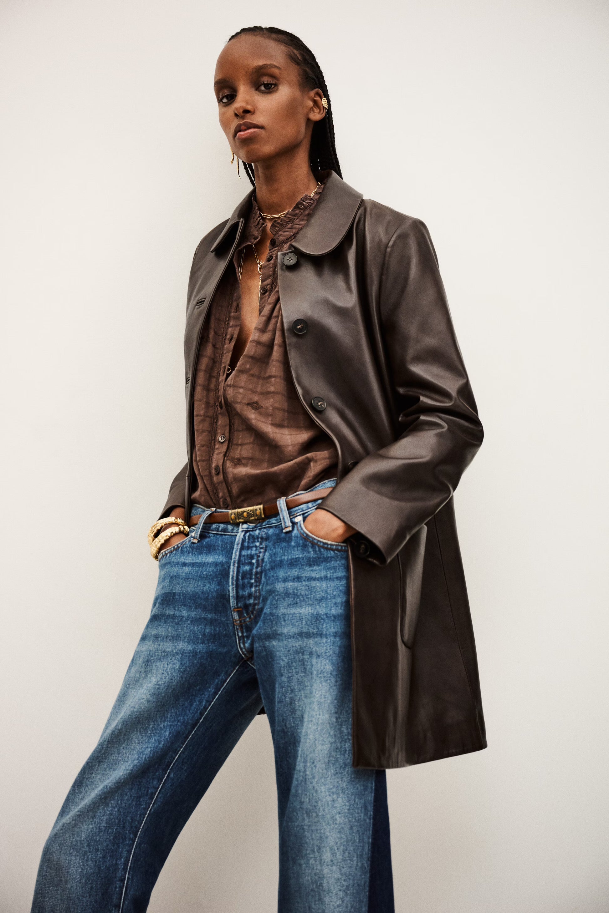 Person wearing a brown leather coat, brown shirt, and blue jeans against a white background