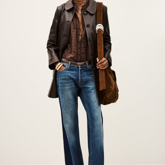 Woman wearing a leather jacket, brown shirt, and blue jeans holding a brown bag on a white background